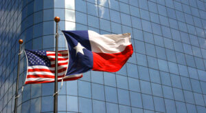 Two flags, USA and Texas, wave against a blue sky with clouds. The backdrop is a modern glass building, creating a sense of patriotism and elegance.
